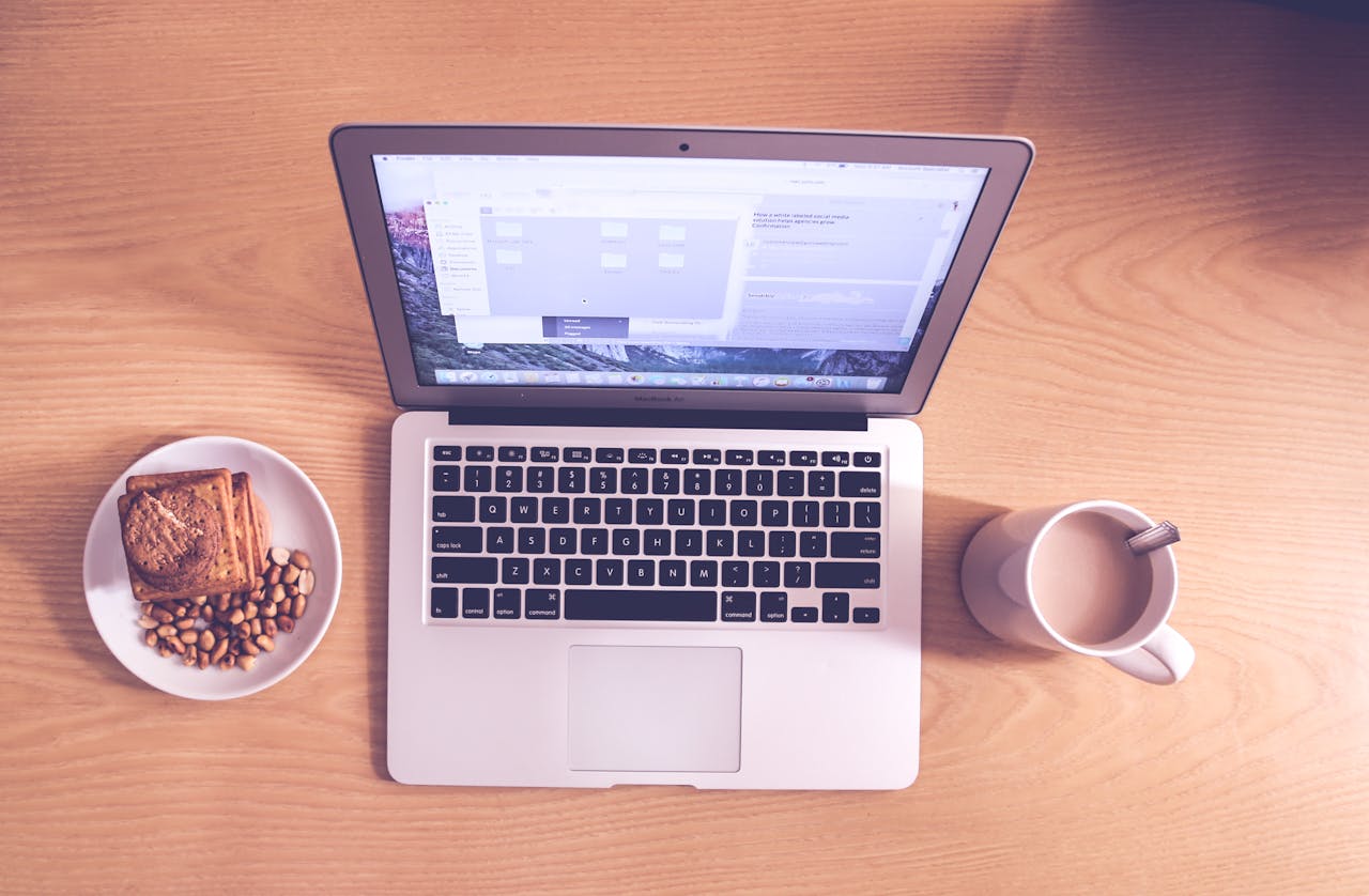 Flat lay of a laptop, coffee mug, and snacks on a wooden desk, ideal for remote work themes.